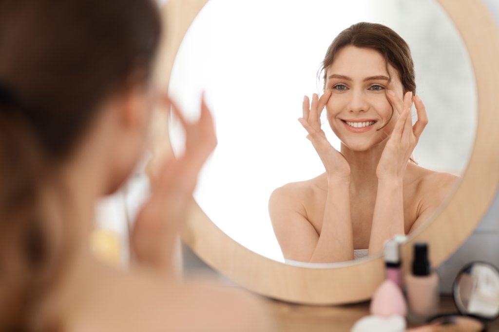 Woman examining her face in mirror with hands near eyes